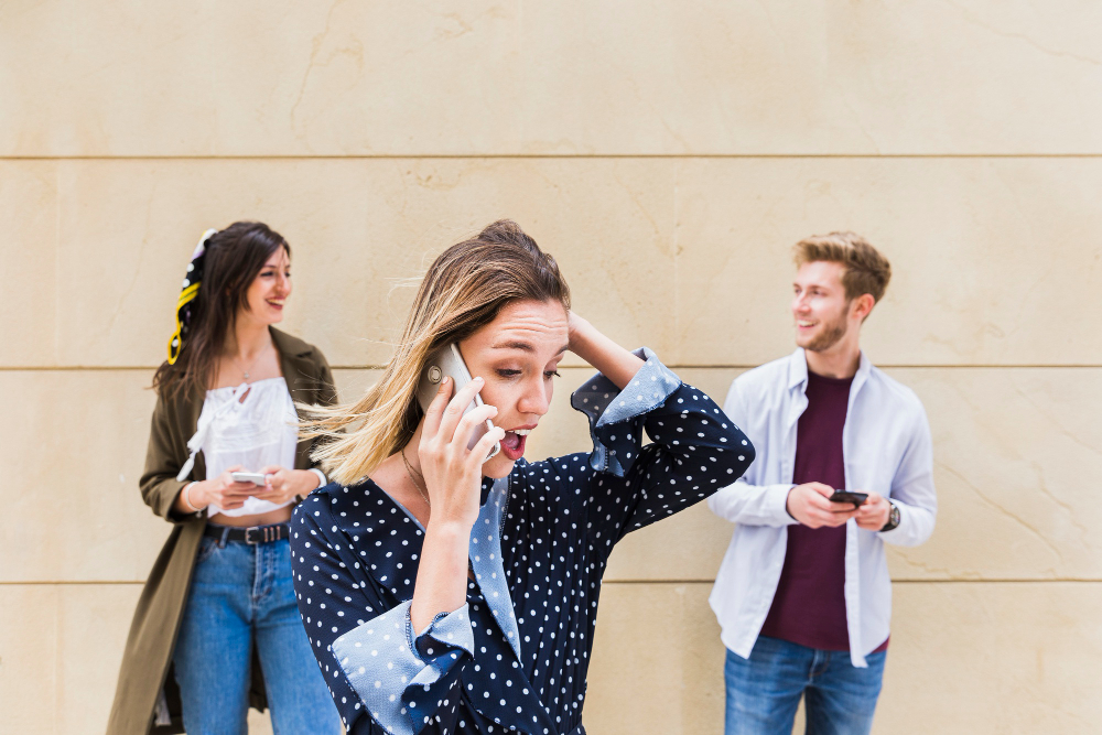 surprised-young-woman-talking-mobile-phone-standing-front-friends-looking-each-other (1)